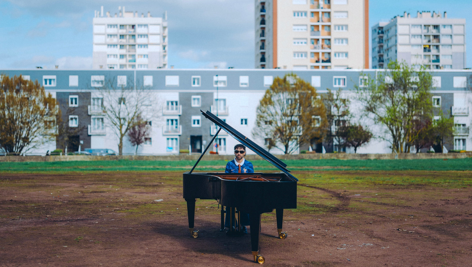 Sofiane Pamart au piano à queue devant des barres d’immeubles, en maillot de l’Équipe de France pour la Coupe du monde