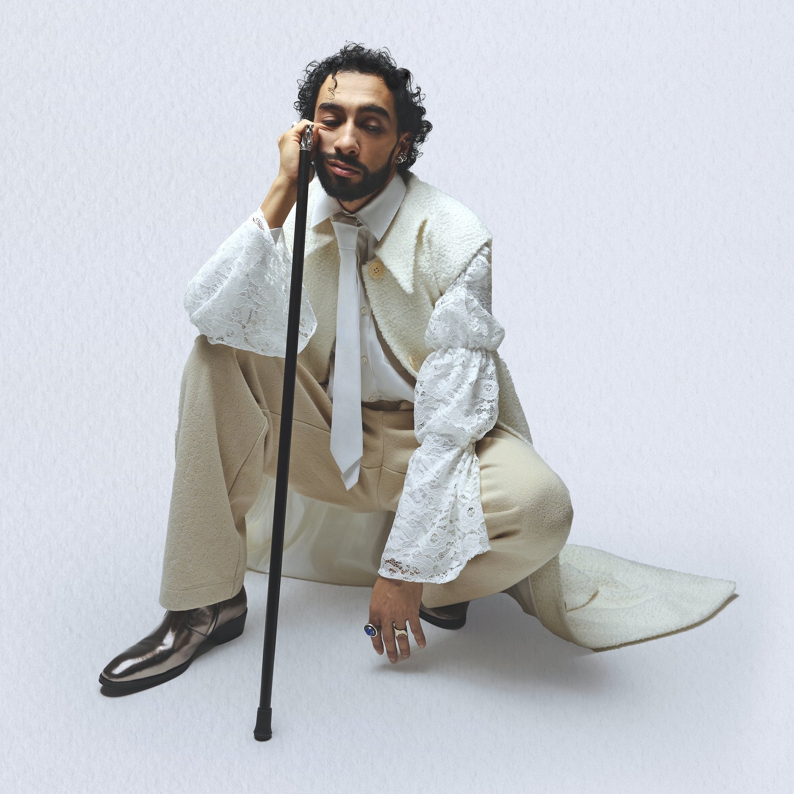 Portrait studio de Sofiane Pamart accroupi en tenue crème, canne à la main
